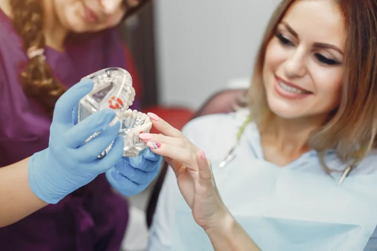 A dentist wearing blue gloves holds a plastic dental model while explaining the procedure to a smiling patient sitting in the dental chair.
