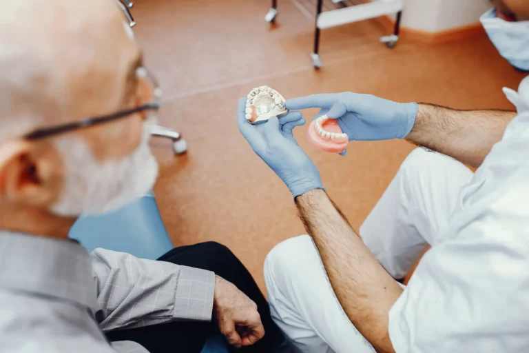 A dentist wearing blue gloves holds and points to dental models while consulting with an elderly male patient.
