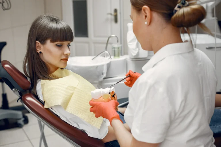 A dentist wearing orange gloves holds a dental model and a small dental tool while consulting with a patient in a dental chair.