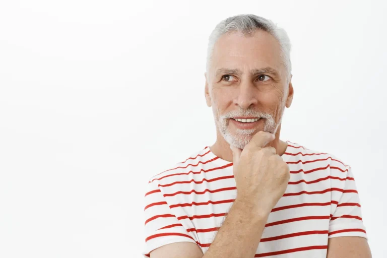 A thoughtful senior man with a gray beard wearing a red and white striped shirt, touching his chin and looking to the side