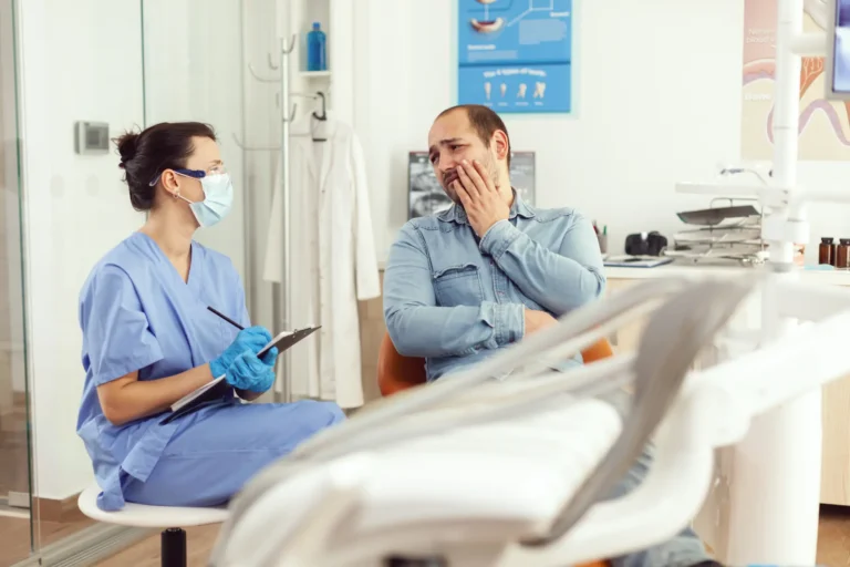 A dentist in protective gear taking notes while a male patient holding his cheek describes his toothache in a dental office.