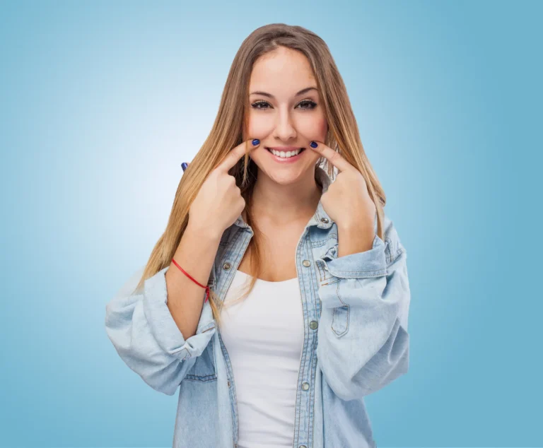 A young woman with long brown hair, wearing an unbuttoned denim shirt over a white top, uses her index fingers to point at and emphasize her smile.