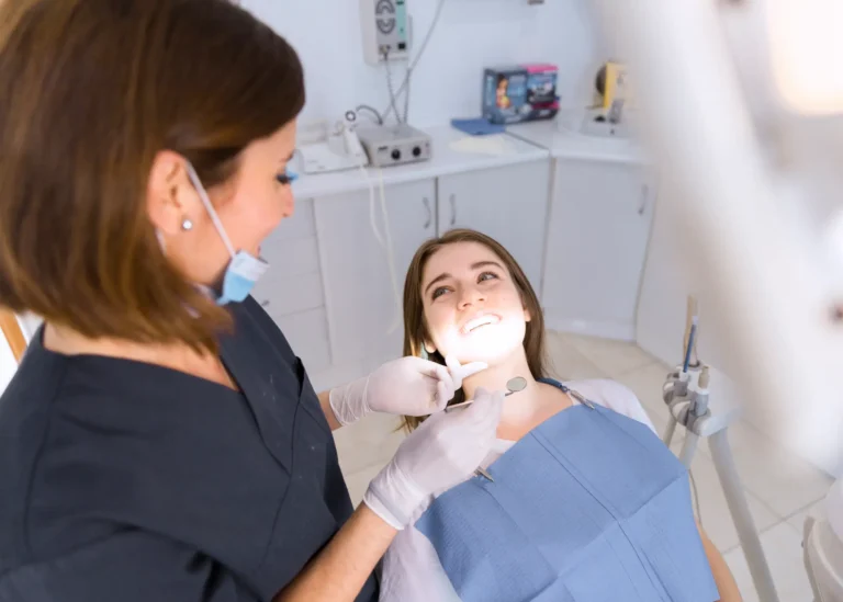 A dentist wearing a mask and gloves using a dental mirror to examine a smiling patient in a dental office